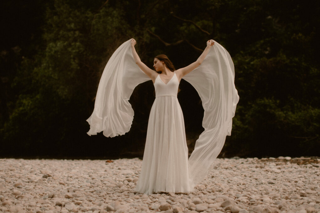 Mariée en robe Atelier Swan à fines bretelles, bras levés, voiles de mousseline flottant au vent